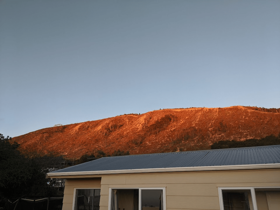 An earth-coloured hill glows orange in the sunset light above a house with a grey steel roof, like a wave of dirt about to swamp the house.