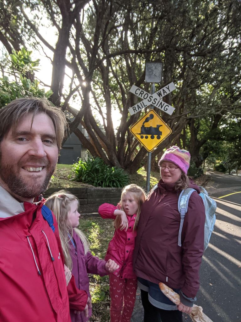 A family poses for a photograph in front of a railway crossing sign. One of the children is coughing.