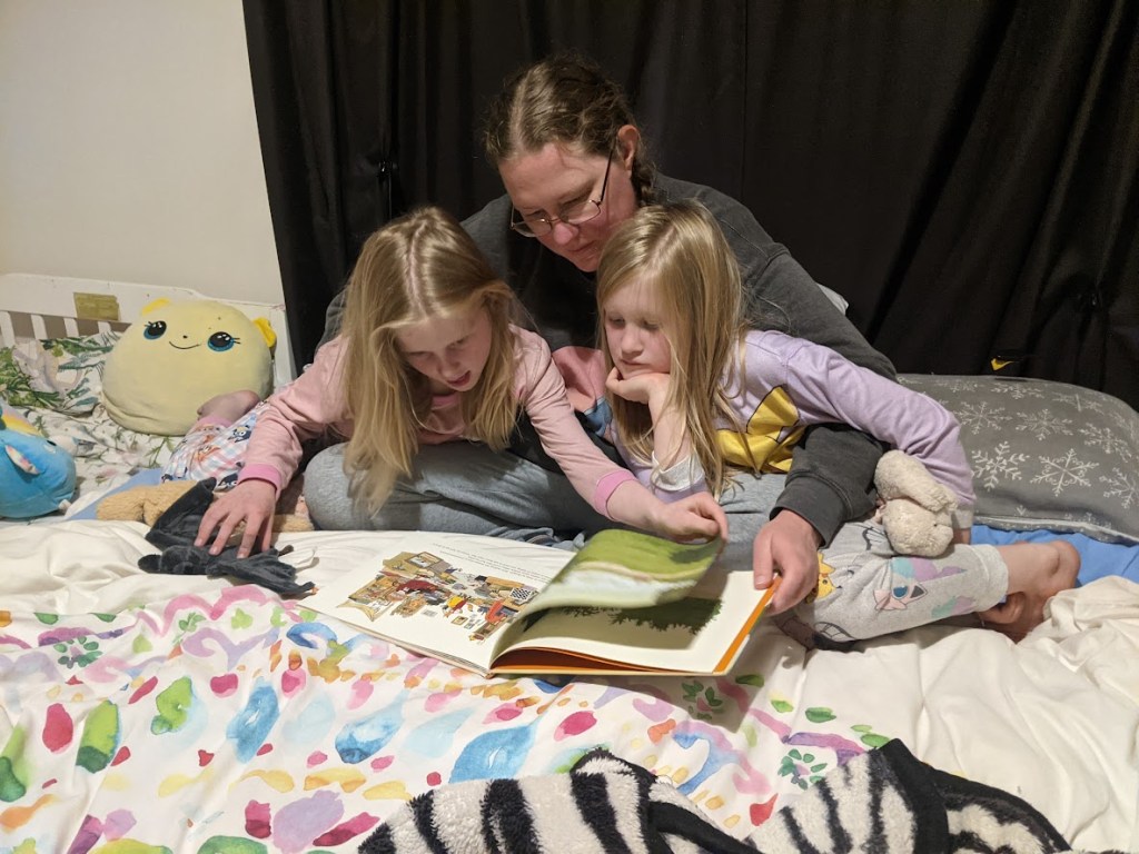 A mother reading a storybook to her two daughters while sitting on a bed, surrounded by colorful bedding and plush toys.