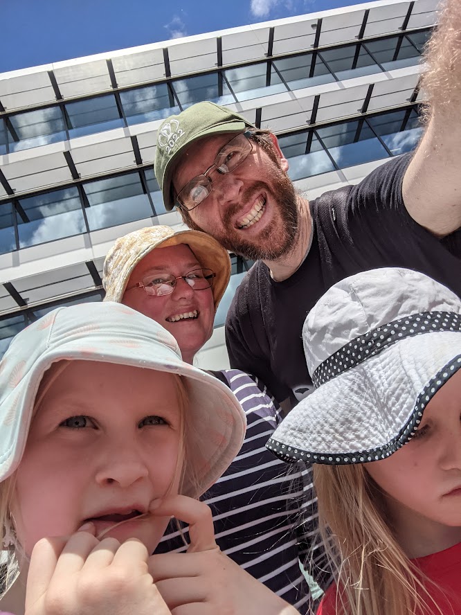 Family selfie with two children and two adults smiling outdoors, wearing hats, in front of a modern building.